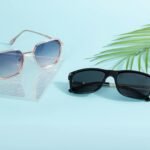 Two pairs of trendy sunglasses on a pastel surface with palm leaf decor.