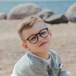 A cheerful young boy wearing glasses sits on a sandy beach under a clear blue sky.